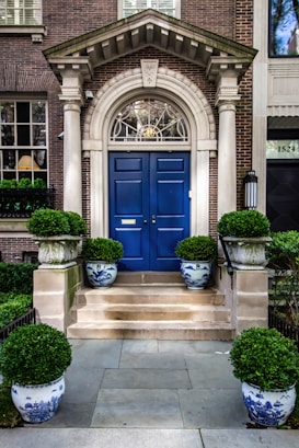 A grand entrance featuring a striking blue double door set within an ornate stone archway. The entrance is accentuated by neatly trimmed topiary plants placed in decorative blue and white porcelain pots. The brick facade of the building provides a classic and elegant backdrop, and a small set of stone stairs leads up to the door.
