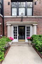The image depicts the entrance to a building, featuring a dark wooden door with glass panels on both sides. Above the door is a decorative lantern. Two large potted plants with pink flowers are placed symmetrically on each side of the entrance, accompanied by trimmed green bushes along the path leading to the door. The building's exterior walls are made of brick, and large windows with curtains can be seen above the doorway.