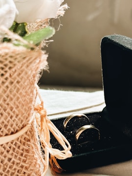 A pair of gold rings is nestled inside an open black velvet box. To the left, a bouquet wrapped in burlap and ribbon features white flowers and green leaves, creating a rustic and elegant composition.