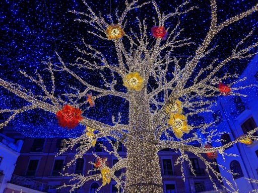 A large artificial tree adorned with countless small white lights stands against a backdrop of deep blue lights, resembling a starry sky. Red and yellow floral decorations are interspersed among the branches, adding vibrant splashes of color. Buildings with balconies are visible in the background, suggesting an urban setting.