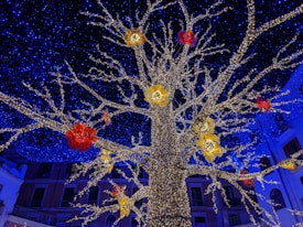 A large artificial tree adorned with countless small white lights stands against a backdrop of deep blue lights, resembling a starry sky. Red and yellow floral decorations are interspersed among the branches, adding vibrant splashes of color. Buildings with balconies are visible in the background, suggesting an urban setting.