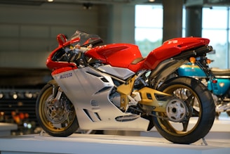 A sleek, red and silver motorcycle on display indoors, featuring a sporty aerodynamic design with prominent fairings and exposed mechanical parts. The bike's frame and swingarm have a gold finish, and the motorcycle rests on a raised platform within a modern exhibition space.