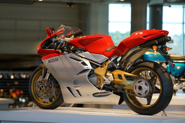 A sleek, red and silver motorcycle on display indoors, featuring a sporty aerodynamic design with prominent fairings and exposed mechanical parts. The bike's frame and swingarm have a gold finish, and the motorcycle rests on a raised platform within a modern exhibition space.