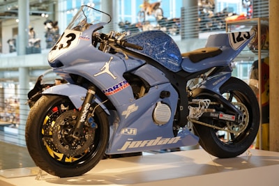 A blue sport motorcycle displayed on a stand in an indoor setting. The motorcycle has various sponsor logos and decals, including a prominent white silhouette of a basketball player and the number 23. The design is sleek and aerodynamic, indicative of a racing bike.