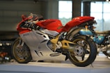A red and silver sports motorcycle is displayed in a well-lit showroom. The sleek design highlights its aerodynamic features and modern engineering. The motorcycle is elevated on a platform, allowing a clear view of its intricate details, such as the wheel rims and the suspension system.