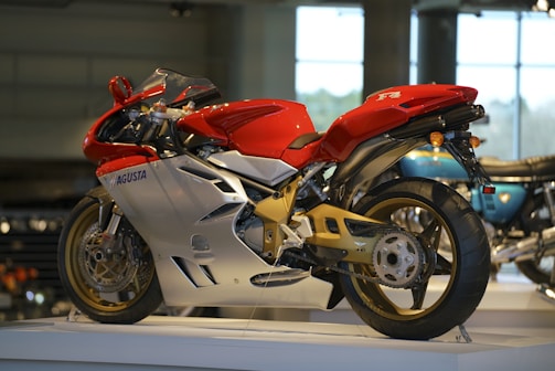 A red and silver sports motorcycle is displayed in a well-lit showroom. The sleek design highlights its aerodynamic features and modern engineering. The motorcycle is elevated on a platform, allowing a clear view of its intricate details, such as the wheel rims and the suspension system.