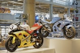 Two racing motorcycles are displayed on a pedestal in a modern showroom. The yellow motorcycle has the number 3 on its front and is adorned with various logos and decals. The blue motorcycle, numbered 25, features sleek design elements and branding. The background includes other motorcycles and polished concrete columns, creating an industrial ambiance with bright overhead lighting.