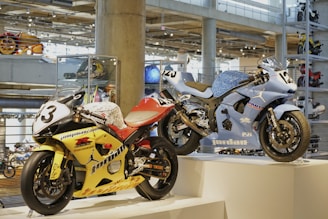 Two racing motorcycles are displayed on a pedestal in a modern showroom. The yellow motorcycle has the number 3 on its front and is adorned with various logos and decals. The blue motorcycle, numbered 25, features sleek design elements and branding. The background includes other motorcycles and polished concrete columns, creating an industrial ambiance with bright overhead lighting.
