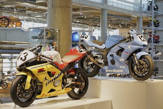Two racing motorcycles are displayed on a pedestal in a modern showroom. The yellow motorcycle has the number 3 on its front and is adorned with various logos and decals. The blue motorcycle, numbered 25, features sleek design elements and branding. The background includes other motorcycles and polished concrete columns, creating an industrial ambiance with bright overhead lighting.