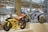 Two racing motorcycles are displayed on a pedestal in a modern showroom. The yellow motorcycle has the number 3 on its front and is adorned with various logos and decals. The blue motorcycle, numbered 25, features sleek design elements and branding. The background includes other motorcycles and polished concrete columns, creating an industrial ambiance with bright overhead lighting.