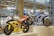 Two racing motorcycles are displayed on a pedestal in a modern showroom. The yellow motorcycle has the number 3 on its front and is adorned with various logos and decals. The blue motorcycle, numbered 25, features sleek design elements and branding. The background includes other motorcycles and polished concrete columns, creating an industrial ambiance with bright overhead lighting.