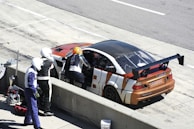 A racing car is parked in a pit stop area with its driver's side door open. Three individuals in racing suits and helmets are standing nearby, one of whom is leaning into the car. The car has a prominent rear wing and a distinctive orange, white, and black color pattern.