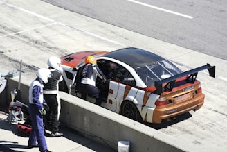 A racing car is parked in a pit stop area with its driver's side door open. Three individuals in racing suits and helmets are standing nearby, one of whom is leaning into the car. The car has a prominent rear wing and a distinctive orange, white, and black color pattern.