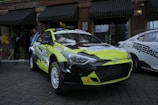 A brightly colored rally car with a yellow and black color scheme is parked on a cobblestone street. The car has various stickers and advertisements on its body, and a Hyundai logo is visible on the front grille. Behind the rally car are several people and another vehicle with a white and black color scheme. The background features a brick building with black awnings and windows, with some graffiti visible on one side.