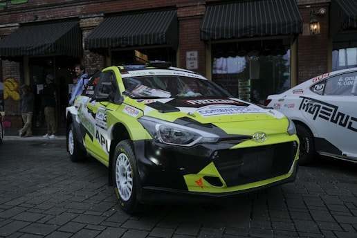 A brightly colored rally car with a yellow and black color scheme is parked on a cobblestone street. The car has various stickers and advertisements on its body, and a Hyundai logo is visible on the front grille. Behind the rally car are several people and another vehicle with a white and black color scheme. The background features a brick building with black awnings and windows, with some graffiti visible on one side.