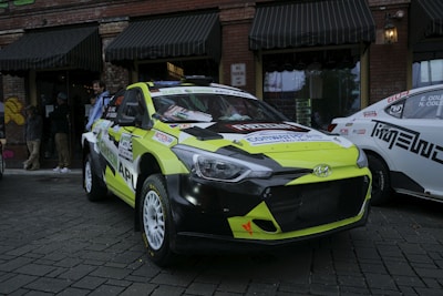A brightly colored rally car with a yellow and black color scheme is parked on a cobblestone street. The car has various stickers and advertisements on its body, and a Hyundai logo is visible on the front grille. Behind the rally car are several people and another vehicle with a white and black color scheme. The background features a brick building with black awnings and windows, with some graffiti visible on one side.