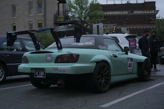 A modified sports car with a large rear spoiler is parked on a street. The car is painted in a light turquoise color and is adorned with various racing stickers and decals, including the number 46 on the side. Two people are standing nearby, dressed in casual clothing. The surroundings include buildings with urban architecture and other parked vehicles, suggesting a city setting.