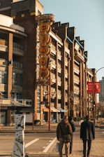 A street view featuring a distinctive vertical 'CHINATOWN' sign on a multi-story brick building. Three people are walking along the sidewalk, and the scene is bathed in a warm, afternoon light. The building has a modern frontage with several balconies and shops at street level.
