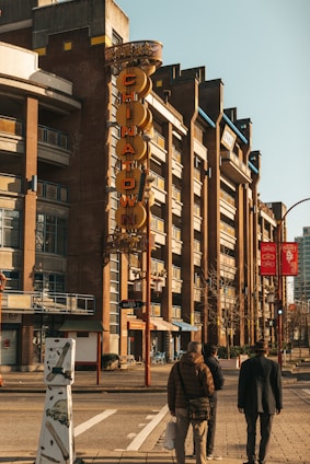A street view featuring a distinctive vertical 'CHINATOWN' sign on a multi-story brick building. Three people are walking along the sidewalk, and the scene is bathed in a warm, afternoon light. The building has a modern frontage with several balconies and shops at street level.