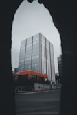 A modern building with a glass facade framed by a dark archway, creating a dramatic perspective. The structure features a distinctive orange corner section on the lower floors, contrasting with the muted sky.