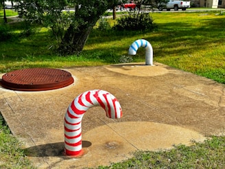 Two curved pipes protrude from a concrete surface; one pipe is painted with red and white stripes, resembling a candy cane, while the other features blue and white stripes. Nearby, a round metal manhole cover is embedded in the ground. The surrounding area is grassy with trees and a road in the background, along with parked white vehicles.