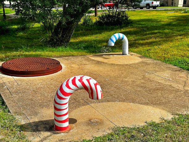 Two curved pipes protrude from a concrete surface; one pipe is painted with red and white stripes, resembling a candy cane, while the other features blue and white stripes. Nearby, a round metal manhole cover is embedded in the ground. The surrounding area is grassy with trees and a road in the background, along with parked white vehicles.