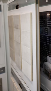A display of various tiles is arrayed vertically. The foreground features light-colored tiles with a subtle texture, arranged in rows, while the background contains darker, glossy tiles with a distinctive geometric pattern.