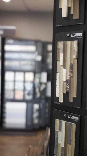 A display of flooring samples is presented on black boards, showcasing a variety of tile and stone options. The samples are organized in straight lines and include different textures and colors such as beige, white, and brown. The foreground focuses on these samples under the company brand Shaw Floors, while a blurred background suggests more displays in a showroom setting.