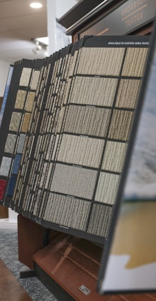 An assortment of carpet samples displayed on a standing rack, featuring various textures and colors including beige, blue, and gray. The setup is indoors, likely in a showroom or retail store, with a focus on home decor options.