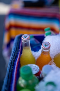 Several colorful bottled drinks are partially submerged in crushed ice inside a cooler, with vibrant stripes of a textile in the background.