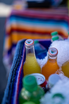 Several colorful bottled drinks are partially submerged in crushed ice inside a cooler, with vibrant stripes of a textile in the background.