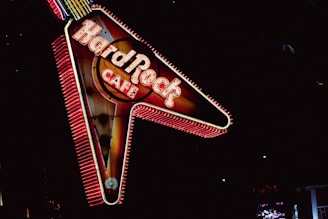 A brightly lit neon sign in the shape of a guitar with the words Hard Rock Cafe. The sign is illuminated with vibrant red and yellow lights against a dark background, creating a striking visual contrast.