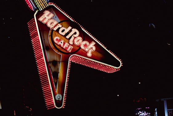 A brightly lit neon sign in the shape of a guitar with the words Hard Rock Cafe. The sign is illuminated with vibrant red and yellow lights against a dark background, creating a striking visual contrast.