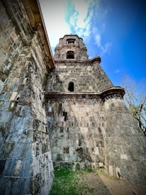 An ancient stone structure with a multi-tiered tower rises against a bright blue sky. The building exhibits weathered stone bricks and intricate architectural details. The view is from a low angle, highlighting the height and grandeur of the tower. Sparse vegetation can be seen at the base, indicating an old historical site.