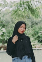 A woman wearing a black hijab and a matching black top stands outdoors in a lush, green environment. She is posing casually with one hand touching her hijab. The background features green foliage and blurred plants, suggesting a natural or garden setting.