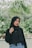 A woman wearing a black hijab and a matching black top stands outdoors in a lush, green environment. She is posing casually with one hand touching her hijab. The background features green foliage and blurred plants, suggesting a natural or garden setting.