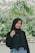 A woman wearing a black hijab and a matching black top stands outdoors in a lush, green environment. She is posing casually with one hand touching her hijab. The background features green foliage and blurred plants, suggesting a natural or garden setting.