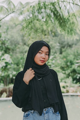 A woman wearing a black hijab and a matching black top stands outdoors in a lush, green environment. She is posing casually with one hand touching her hijab. The background features green foliage and blurred plants, suggesting a natural or garden setting.