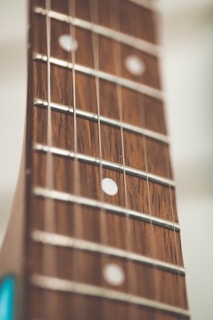 Close-up shot of a guitar fretboard with visible strings and fret markers. The wood grain is detailed and the image has a shallow depth of field, creating a soft, blurred background.
