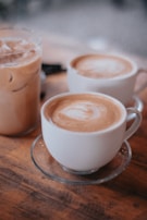Two cups of hot coffee, each with frothy milk designs on top, sit on a wooden table next to a glass of iced coffee. The setting conveys a cozy café atmosphere.