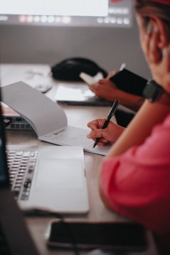 Two people are seated at a table, taking notes with notebooks and pens. A laptop and a smartphone are also present on the table. The atmosphere seems focused and collaborative.