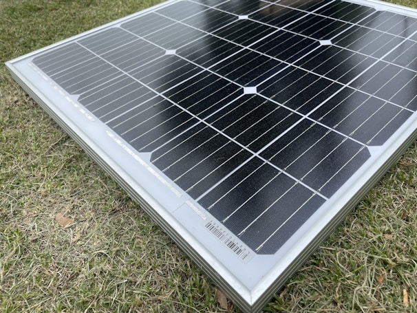 A solar panel resting on a grass surface with visible grass blades surrounding the panel. The solar panel has a grid of black photovoltaic cells separated by white lines.