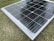 A solar panel resting on a grass surface with visible grass blades surrounding the panel. The solar panel has a grid of black photovoltaic cells separated by white lines.