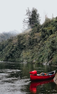 A serene river scene with a bright red canoe gently floating on the calm water, surrounded by lush, dense greenery of a forested hillside. The background consists of tall trees shrouded in light mist, creating a tranquil atmosphere.