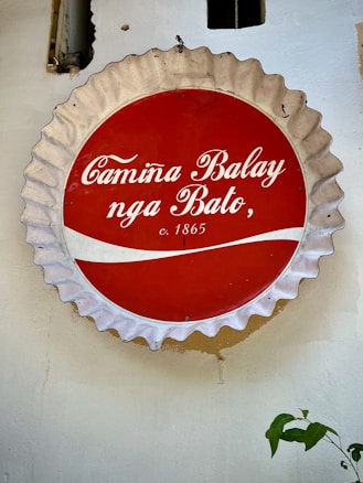 A circular vintage sign resembling a bottle cap is mounted on a wall. The sign has a red center with white cursive letters reading 'Camina Balay nga Bato, c. 1865.' The wavy white pattern runs through the red section. The border of the sign is a metallic silver color. Part of a plant with green leaves is visible at the bottom right corner.