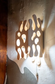 A uniquely designed wall light fixture with an abstract, wavy wooden panel. The panel has numerous cut-out holes allowing light to pass through, creating a warm and artistic display on the textured wall. The wood has a natural finish and the fixture is attached by a visible wire.