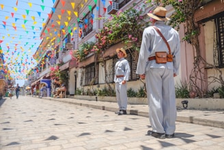 A scene on a cobblestone street adorned with colorful triangular flags hanging above. Two people wearing wide-brimmed hats and traditional clothing stand facing each other. The street is lined with colonial-style buildings, and vibrant flowers cascade from balconies. Other people and a bicycle are visible further down the street, adding to the lively atmosphere.
