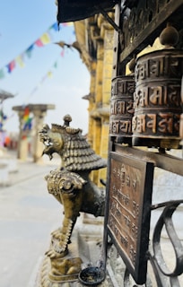 A bronze or metallic prayer wheel with inscriptions in a temple setting is prominently displayed. Nearby, a statue resembling a mythical lion-like creature adds to the cultural ambiance. Colorful prayer flags are blurred in the background, enhancing the spiritual atmosphere.