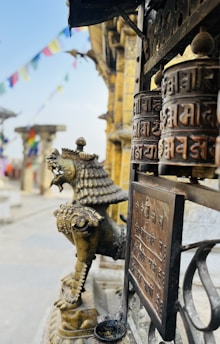 A bronze or metallic prayer wheel with inscriptions in a temple setting is prominently displayed. Nearby, a statue resembling a mythical lion-like creature adds to the cultural ambiance. Colorful prayer flags are blurred in the background, enhancing the spiritual atmosphere.