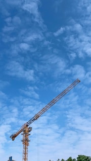 A large construction crane is prominently featured against a bright blue sky with scattered, wispy clouds. The crane is tall and yellow, stretching across the sky. Below the crane, there is a hint of buildings and treetops.